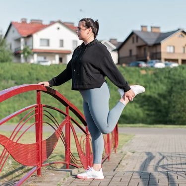 A woman in active wear performs a stretching exercise on a red bridge in a suburban park setting.